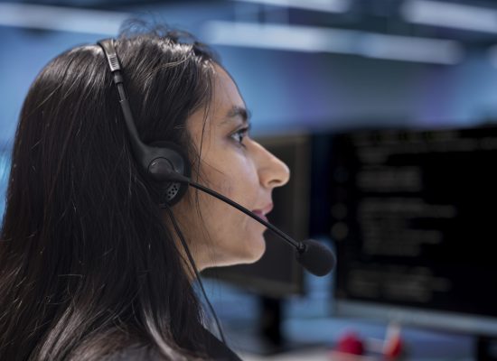 Admin in data center facility managing call center tasks, speaking into headset microphone while monitoring multiple PC screens. Indian woman talking with customers through headphones mic