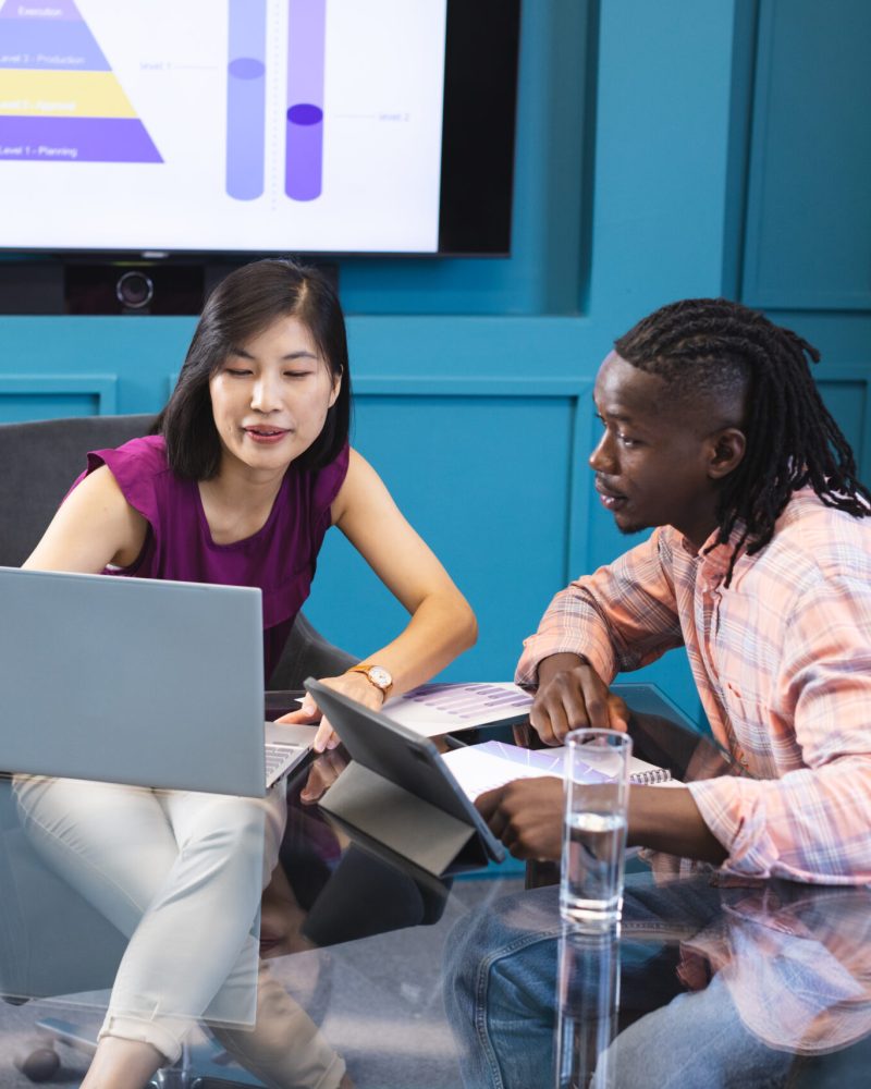 Asian woman and African American man looking at laptop screen together in a modern business office. She has black hair, he wears a checkered shirt, both young and focused, unaltered.