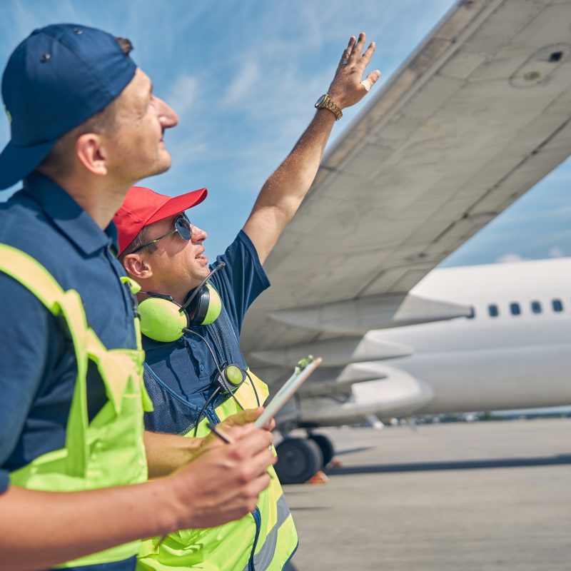 Side view of a professional maintenance engineer pointing at an airliner wing to his colleague