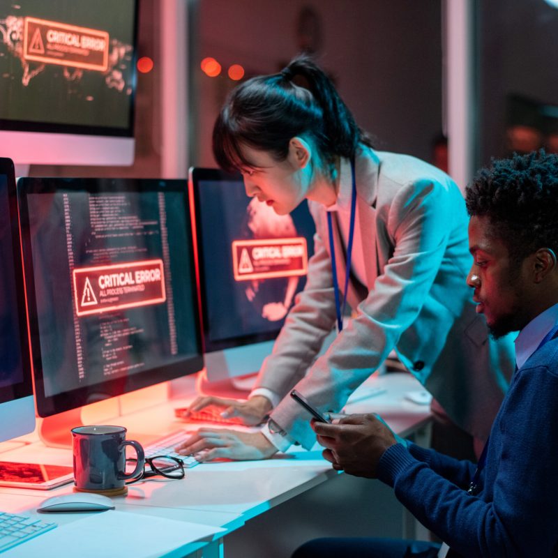 Two young intercultural programmers trying to solve technical problem in front of computers with critical error on screens