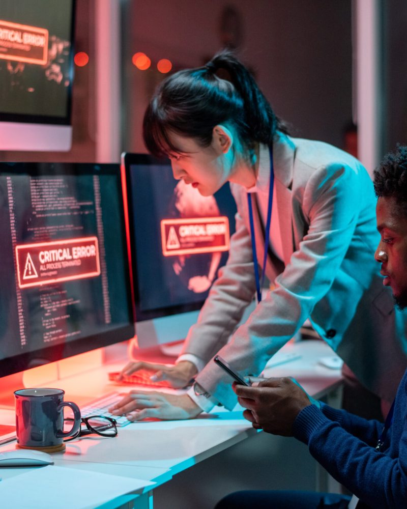 Two young intercultural programmers trying to solve technical problem in front of computers with critical error on screens